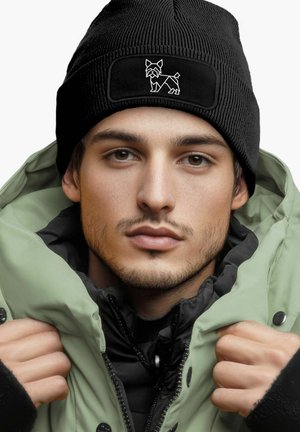 Black ribbed beanie with a textured surface and a rectangular patch featuring a white dog graphic. Worn with a green puffer jacket.