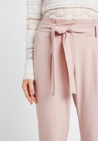 Femme portant un pantalon taille haute rose clair avec une ceinture nouée et un haut blanc en dentelle à manches longues, main détendue le long du corps.