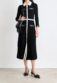 Robe plissée noire avec un col blanc et une fermeture à boutons sur le devant, ornée de rayures blanches d écoratives et de deux poches. Accompagnée de chaussures à talons ouverts noires.