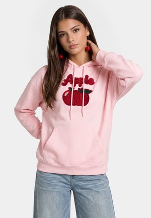 Giovane donna con lunghi capelli castani indossa una felpa con cappuccio rosa con design di mela rossa e orecchini a forma di ciliegia rossa, abbinati a jeans azzurri chiari.