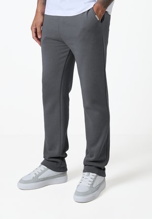 Homme portant un pantalon décontracté gris foncé et des baskets blanches, debout avec une main dans la poche devant un fond blanc uni.