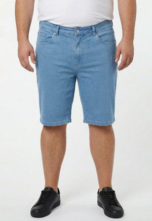 Mand iført lyseblå denimshorts, hvid skjorte og sorte sneakers står mod en ensfarvet hvid baggrund.