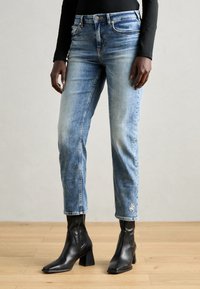 Hellblaue Jeans aus Denim mit Slim-Fit, leichtem Verblassen und gesticktem Ankerdetail in der Nähe des Saums. Kombiniert mit schwarzen Ankle-Boots.
