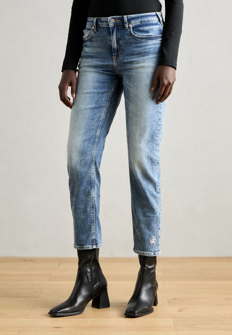 Hellblaue Jeans aus Denim mit Slim-Fit, leichtem Verblassen und gesticktem Ankerdetail in der Nähe des Saums. Kombiniert mit schwarzen Ankle-Boots.