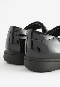 Zapatos de charol negro con una superficie suave, forma redondeada y suelas acolchadas. Presentan un tacón bajo y una correa en la parte trasera para mayor soporte.