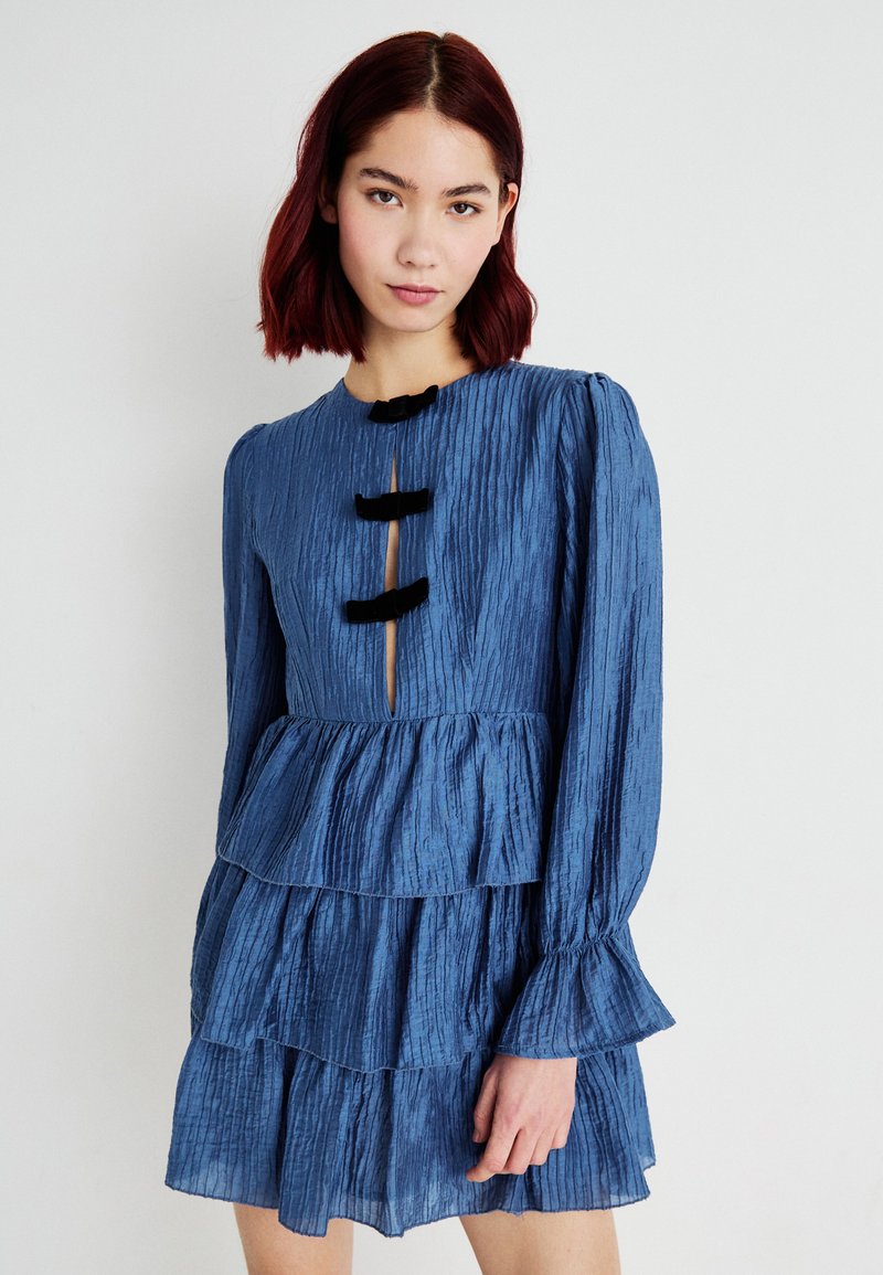 Vestido azul plissado com três laços pretos, mangas longas e um design em camadas com uma abertura em forma de chave na gola.