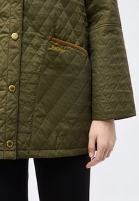 Πράσινο χακί καπιτονέ μπουφάν με μοτίβο διαμαντιού, τσέπη με καφέ λεπτομέρεια κεντημένη με τη λέξη "Barbour" και χρυσά κουμπιά snap, φορεμένο με μαύρο παντελόνι.