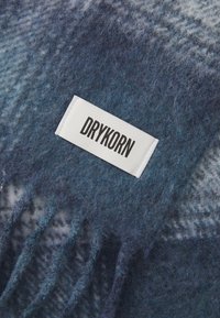 Blå og grå frynset uldhalstørklæde med et hvidt rektangulært mærke syet på, som viser teksten "DRYKORN" i sorte bogstaver.