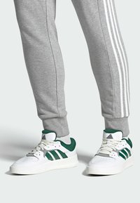 White athletic shoes with green accents, three green stripes, and a textured sole, paired with gray sweatpants featuring white side stripes.