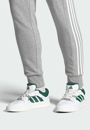 Weiße Sportschuhe mit grünen Akzenten, drei grünen Streifen und einer strukturierten Sohle, kombiniert mit grauen Jogginghosen, die weiße Seitenstreifen haben.