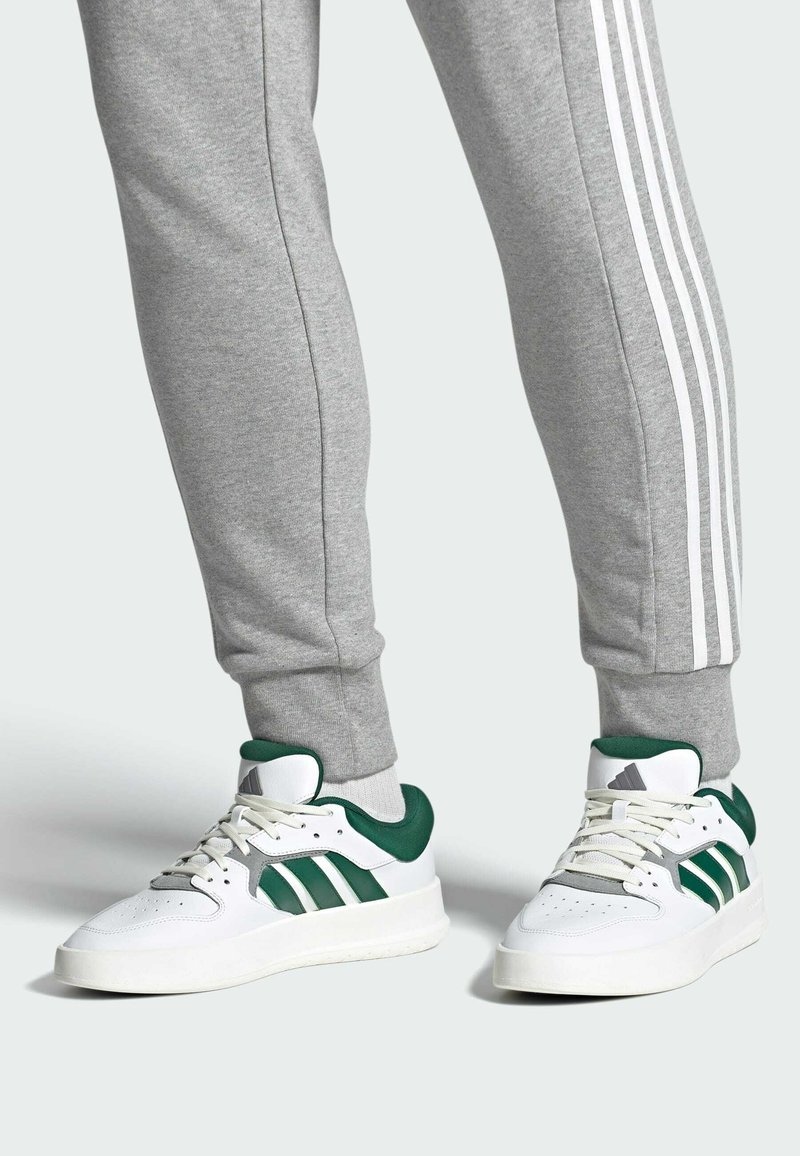 White athletic shoes with green accents, three green stripes, and a textured sole, paired with gray sweatpants featuring white side stripes.