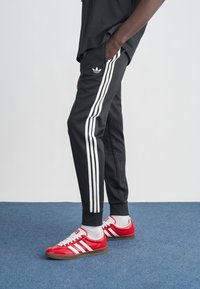 Čierne teplákové nohavice Adidas s bielym dizajnom so tromi pruhmi, v kombinácii so červenobielymi teniskami Adidas, nosené s sivými ponožkami na modrom koberci.