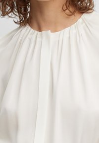 Biała bluzka z plisowanym dekoltem, miękka, lekka tkanina, z pionowym zapięciem z przodu, gładka tekstura, minimalistyczny design.