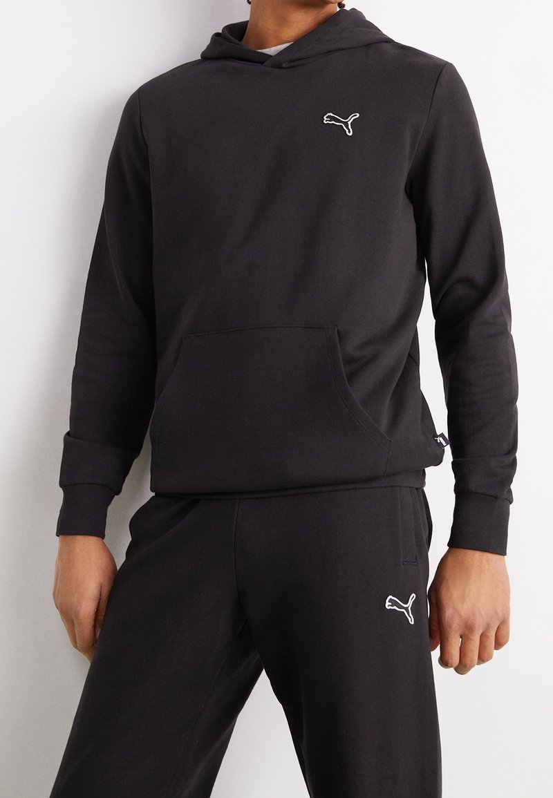 Homme portant un sweat à capuche noir Puma et un pantalon de survêtement noir assorti, tous deux avec le logo blanc Puma sur la poitrine et la cuisse.