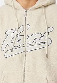Giacca zip-up con cappuccio color crema realizzata in pile morbido, caratterizzata da un logo prominente ricamato in bianco e nero, con lacci e tirazip.
