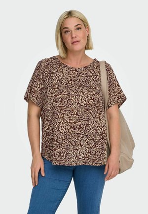 Kvinde med kort blondt hår iført en brun og beige mønstret kortærmet top og blå jeans, bærer en beige skuldertaske.