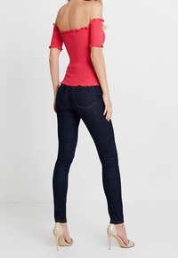 Off-shoulder, smocket, lyserød top med flæsedetaljer, parret med mørkeblå tætsiddende jeans og nude højhælede sko.