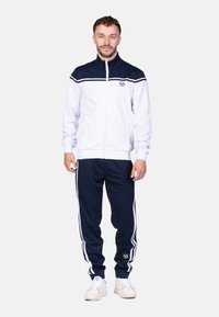 Heren tracksuit met een witte ritsjack met een marineblauwe kraag en strepen. Marineblauwe broek met witte zijstrepen. Stof lijkt glad te zijn.