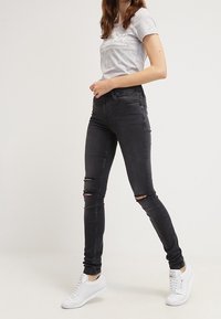 Kvinna som bär en grå grafisk t-shirt, svarta slitna skinny jeans och vita sneakers, stående med ett ben framåt mot en enfärgad bakgrund.