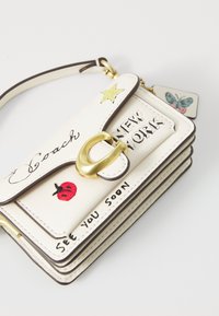Witte leren crossbody tas met gouden hardware, voorzien van geborduurde accenten van een lieveheersbeestje, ster en vlinder. Tekst leest "Coach" en "NEW YORK."