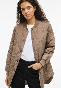 Ruda dygsniuota bombonešio striukė su ribinėmis rankogaliais ir apykakle, su spaudžiamais užsegimais. Dėvima ant balto marškinio ir juodų kelnių.