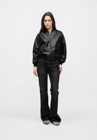 Black leather bomber jacket with a front zip, paired with flared dark denim jeans. Model stands on a plain white background.