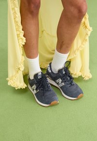 Sneakers gri din nubuc cu tălpi albe și accente cu model. Asortate cu șosete albe până la gleznă și o rochie galbenă cu volane. Fundal texturat verde.