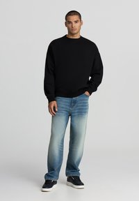 Felpa nera a collo rotondo abbinata a jeans dritti in denim chiaro. Il modello indossa sneakers nere. Un outfit semplice e informale.