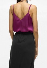 Haut en satin à col en V de couleur magenta vif avec de fines bretelles, associé à une jupe noire à taille élastique. Texture lisse et design fluide visibles.