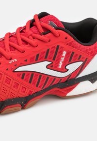 Zapatilla deportiva roja con parte superior de malla, acentos negros y logotipo blanco. Presenta una superficie texturizada, cordones prominentes, diseño deportivo y suela de goma.