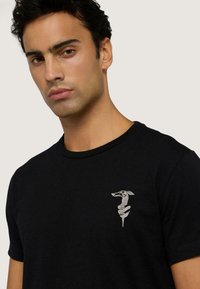 Czarny bawełniany t-shirt z krótkim rękawem, z małą haftowaną grafiką stylizowanego psa na lewej piersi. Gładka tekstura.