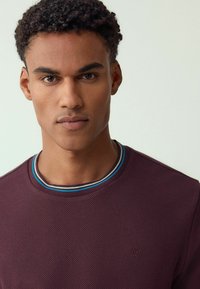 Vyniotas tamsiai raudonas džemperis su apvalia iškirpte, su įvairiaspalviu dryžuotu apvadais. Mažas logotipo elementas ant krūtinės.