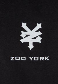 Juodas medvilnės audinys pasižymi ryškiu baltu grafiniu logotipu, primenančiu kampuotus kaulus, su užrašu "ZOO YORK" ryškiu, didžiosiomis raidėmis parašytu šriftu.