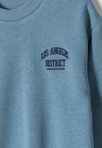 Ljusblå sweatshirt med ribbad halsringning och broderad text "LOS ANGELES DISTRICT" i mörkblått. Mjuk, texturerad tyg.