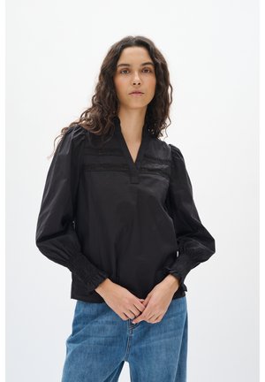 Black long-sleeve blouse with a V-neck, decorative front pleats, and smocked cuffs, paired with blue denim jeans.