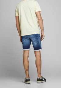 Hombre con pantalones cortos de mezclilla azul enrollados, camiseta de color amarillo pálido, zapatillas y un pequeño tatuaje en la parte posterior de su pantorrilla izquierda, de pie y mirando hacia el lado opuesto.