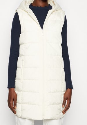 Veste sans manches blanche matelassée à capuche avec fermeture éclair à l'avant, portée sur un haut à manches longues bleu marine foncé et un pantalon blanc.