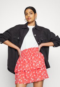 Camisa oversized de denim preto, sobre uma t-shirt branca, combinada com uma saia vermelha em camadas com pequenos padrões florais brancos.