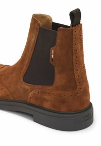 Kahvin mokkanahka Chelsea-saapas mustilla elastisilla sivupaneeleilla, koristeellisella brogue-yksityiskohdalla, vetolenkillä ja kontrastoivalla kumipohjalla.
