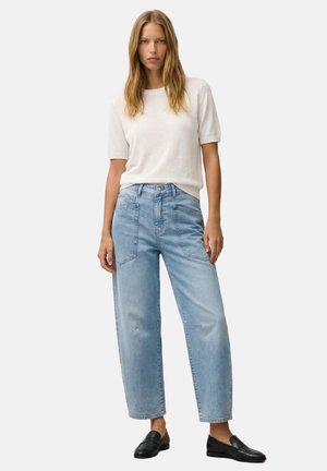 Frau steht vor einfarbigem Hintergrund, trägt hellblaue weite Jeans, ein weißes Kurzarm-Top und schwarze Loafer, schaut nach vorn.