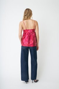 Kvinne iført skinnende rød ermeløs topp med tynne stropper og mørkeblå vide jeans med små hvite prikker, stående foran hvit bakgrunn.