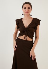 Crop top marrone a costine con maniche corte a volant, scollatura a V profonda, dettaglio con nodo frontale, abbinato a una gonna a vita alta coordinata.