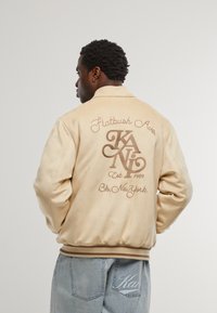 Bež satenski bomber s rebrastim rubom, sa vezom teksta i logotipa na stražnjoj strani. Uparen s svijetloplavim trapericama.