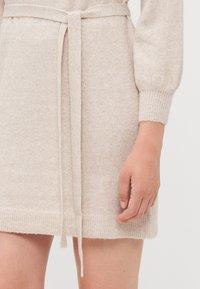 Strikket kjole i beige med teksturert overflate, med belte tål i midjen og ribbestrikkede mansjetter, som slutter over kneet.