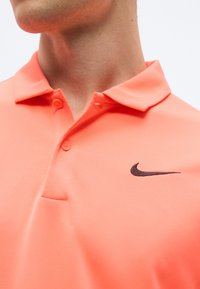 Le cou et le menton d'un homme sont visibles, il porte un polo Nike corail avec deux boutons et un logo swoosh noir sur la poitrine droite.