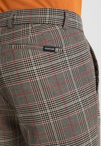 Pantalones de cuadros en gris, negro y rojo, con un tejido texturizado y un bolsillo trasero con una etiqueta "YOURTURN". Diseño de patrón de cuadros clásico.