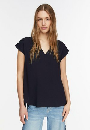 Młoda kobieta o długich falowanych włosach, ubrana w czarną koszulkę z krótkim rękawem i dekoltem w serek oraz jasnoniebieskie dżinsy, stojąca na tle jednolitego białego tła.