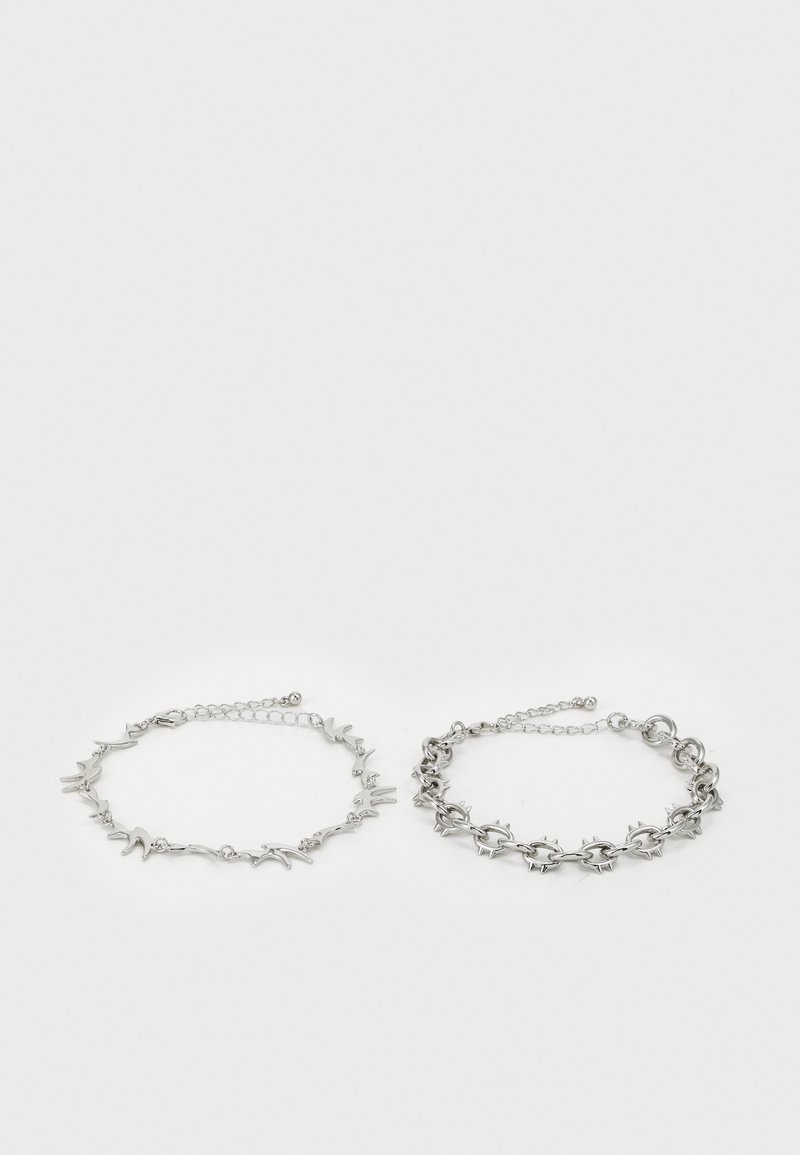 Dos pulseras de plata exhibidas una al lado de la otra. Una presenta una delicada cadena con acentos en forma de pájaro; la otra tiene un diseño audaz y con picos.