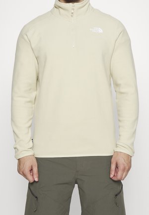 Homme portant un pull polaire beige à fermeture éclair 1/4 avec le logo The North Face et un pantalon vert olive avec des poches zippées.