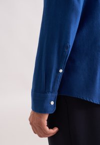 Camicia blu a maniche lunghe con polsini abbottonati, realizzata in un tessuto morbido. Presenta una finitura leggermente testurizzata e un piccolo logo stampato vicino alla manica.